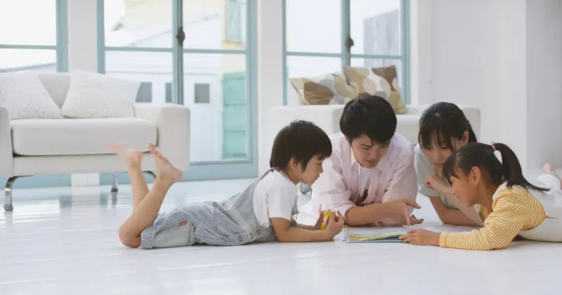 リビングの床に寝転がりながら本を読む親子4人の写真